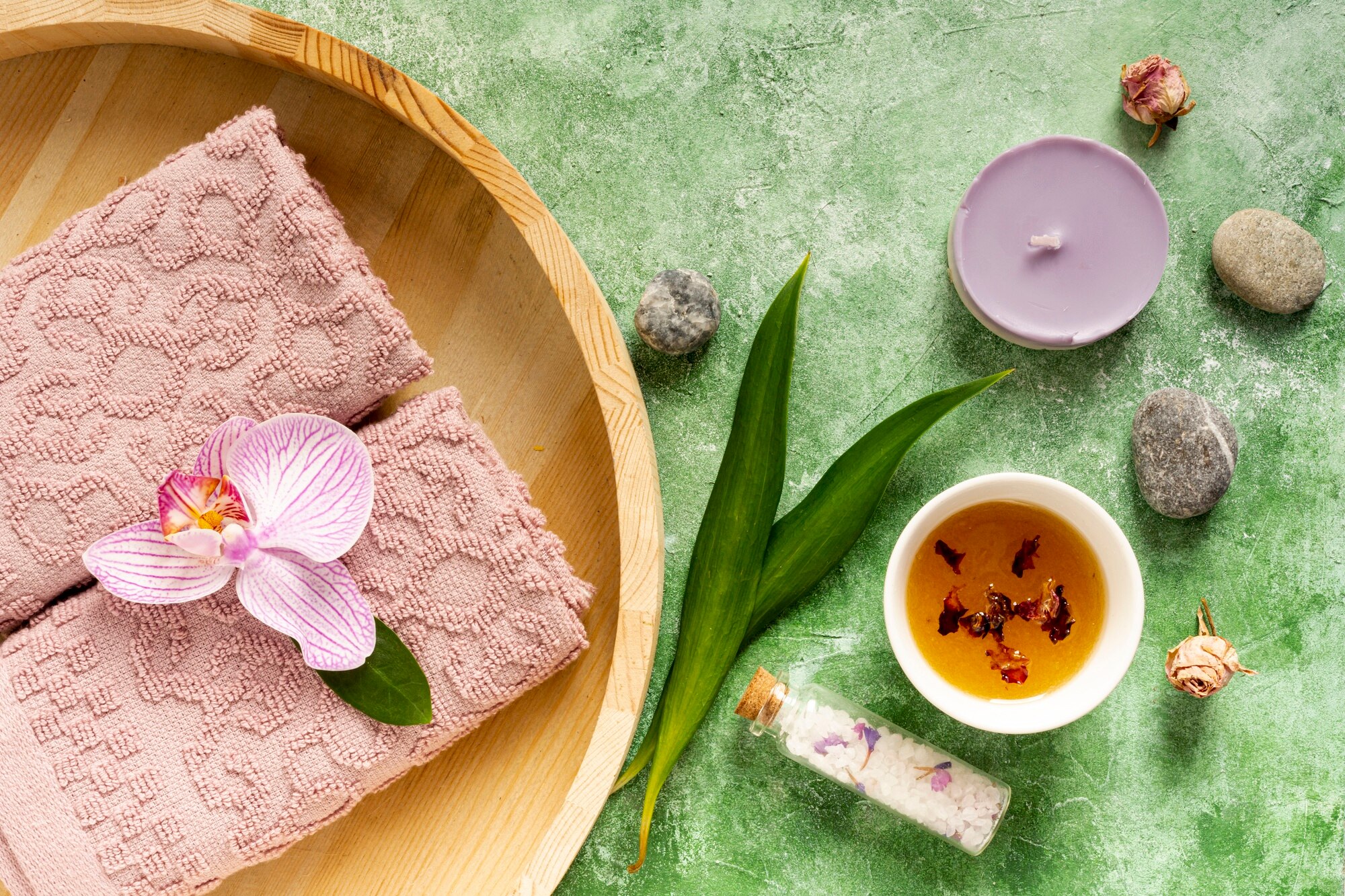 Soft pink towels are arranged with an orchid, essential oils, and a candle, creating a calming spa-like atmosphere on a green textured background.
